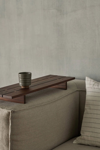 The ferm LIVING Serena Cup with a reactive glaze rests on a wooden tray atop a beige sofa’s armrest, beside a light striped cushion. A plain, light gray wall forms the background.