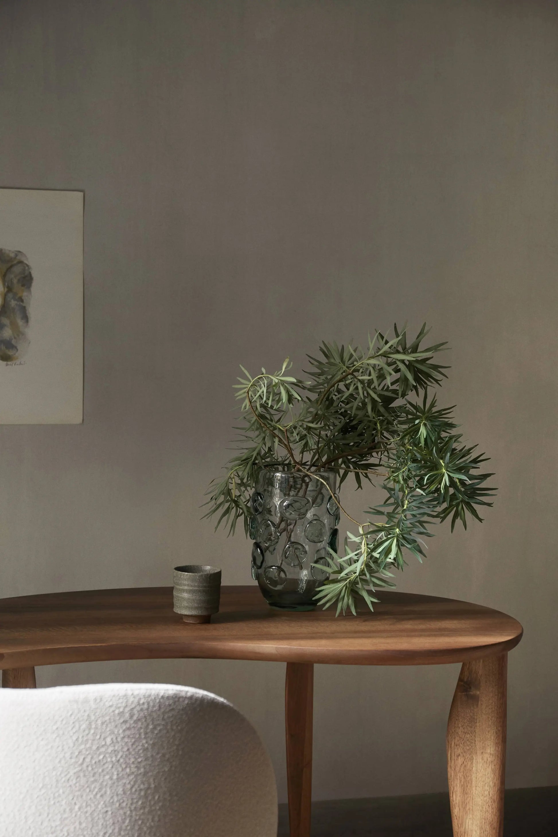 A wooden table holds a green leafy branch in a textured glass vase and the ferm LIVING Serena Cup. A light chair and abstract wall art are partly visible in this softly lit, minimalist room.