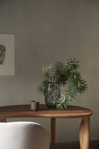 A wooden table holds a green leafy branch in a textured glass vase and the ferm LIVING Serena Cup. A light chair and abstract wall art are partly visible in this softly lit, minimalist room.