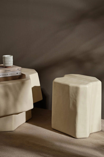A minimalist scene with a beige, irregular stool beside a matching side table holding stacked books and the ferm LIVING Serena Cup, set against a taupe wall with soft shadows.