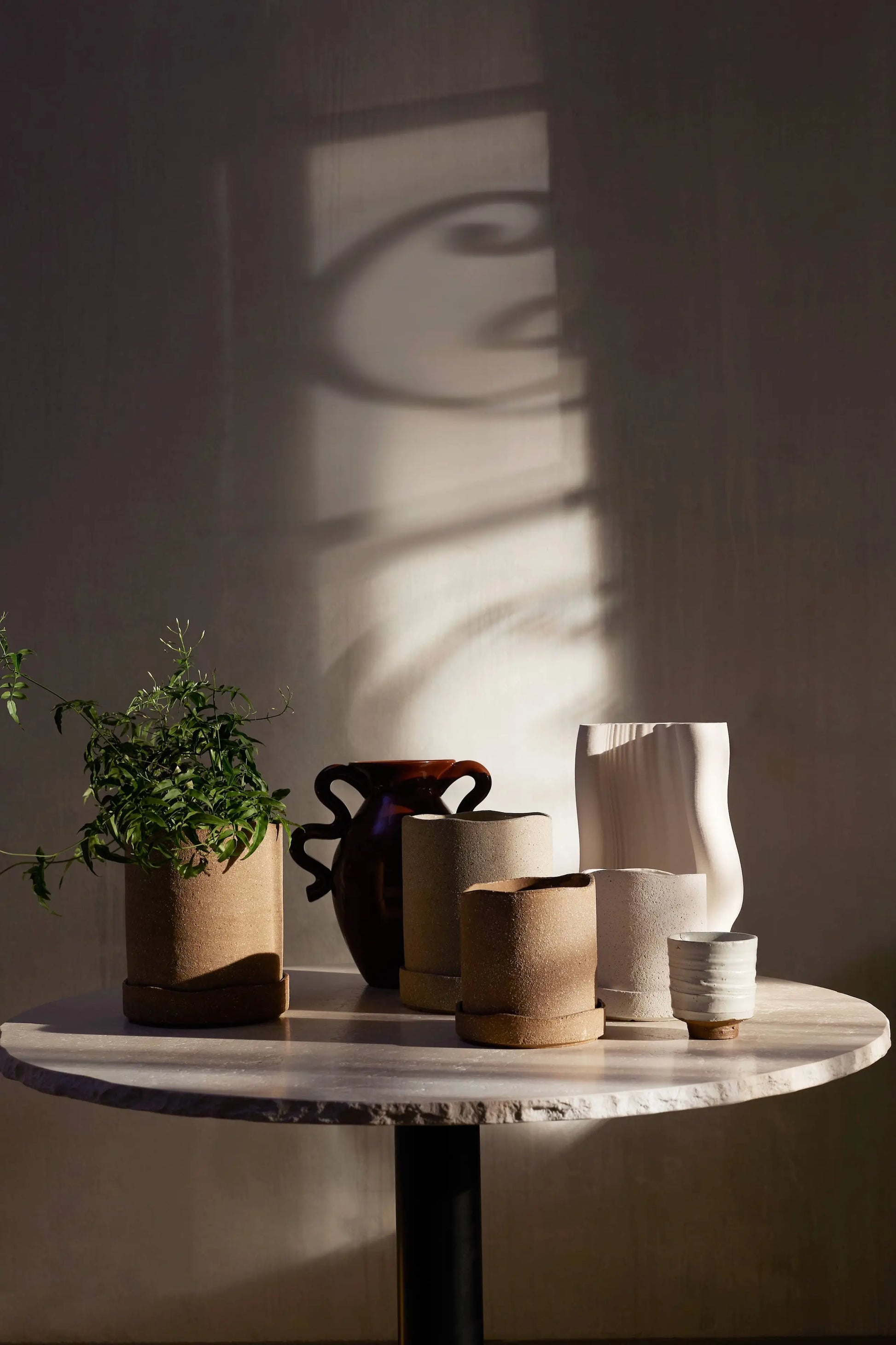 A round table features the ferm LIVING Serena Cup, pottery vases, a plant in a brown pot, and a ribbed white vase. Sunlight and shadows cast abstract patterns on the wall in the background.