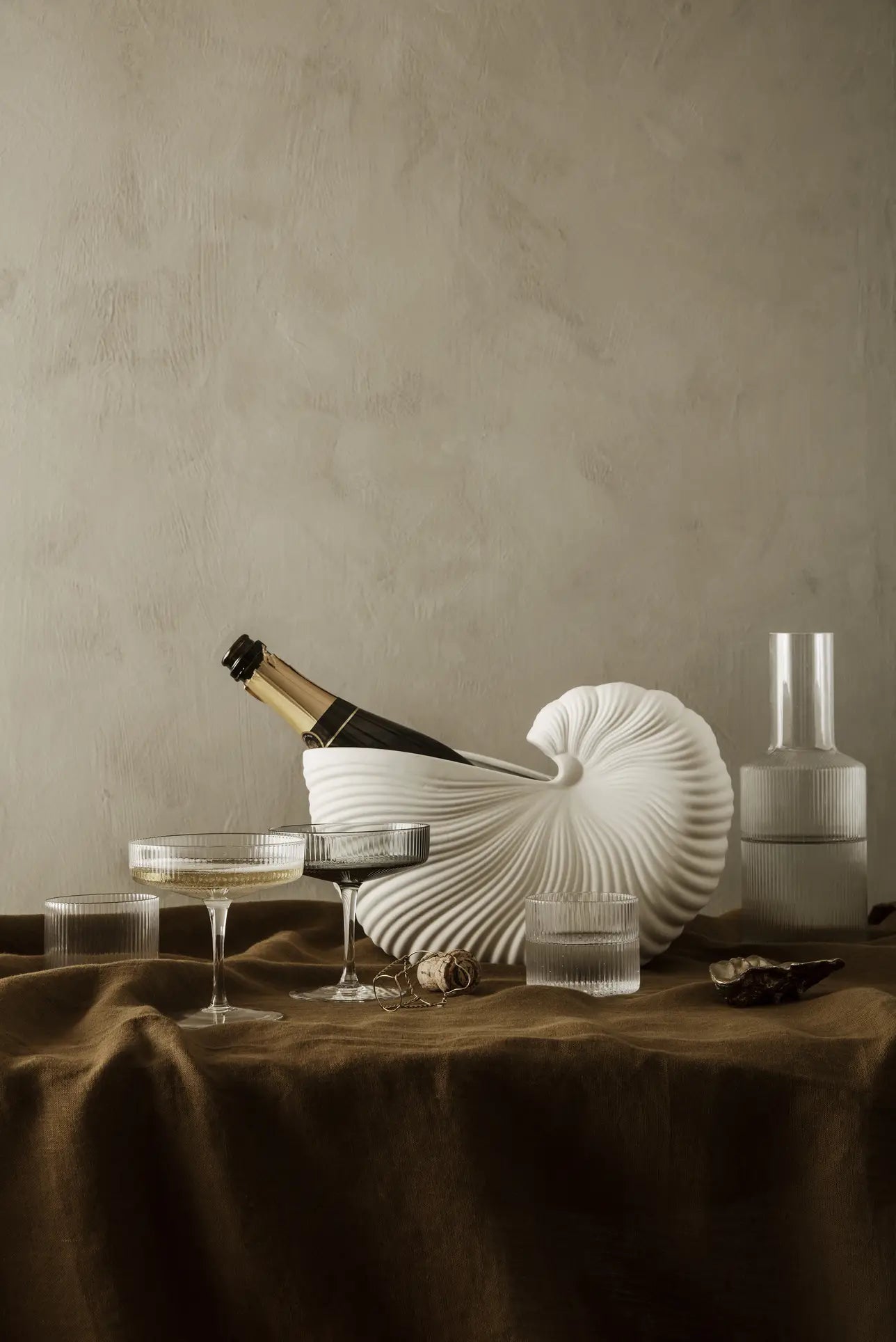 A champagne bottle sits in the ferm LIVING Shell Pot by ferm LIVING on a brown tablecloth, surrounded by two coupe glasses, two short glasses, a cork, and a clear carafe against a neutral textured wall—a nod to modern design.