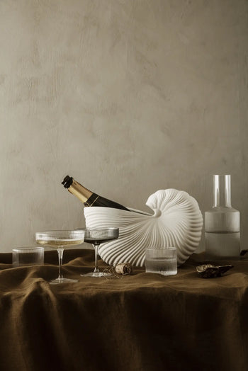 A champagne bottle sits in the ferm LIVING Shell Pot by ferm LIVING on a brown tablecloth, surrounded by two coupe glasses, two short glasses, a cork, and a clear carafe against a neutral textured wall—a nod to modern design.