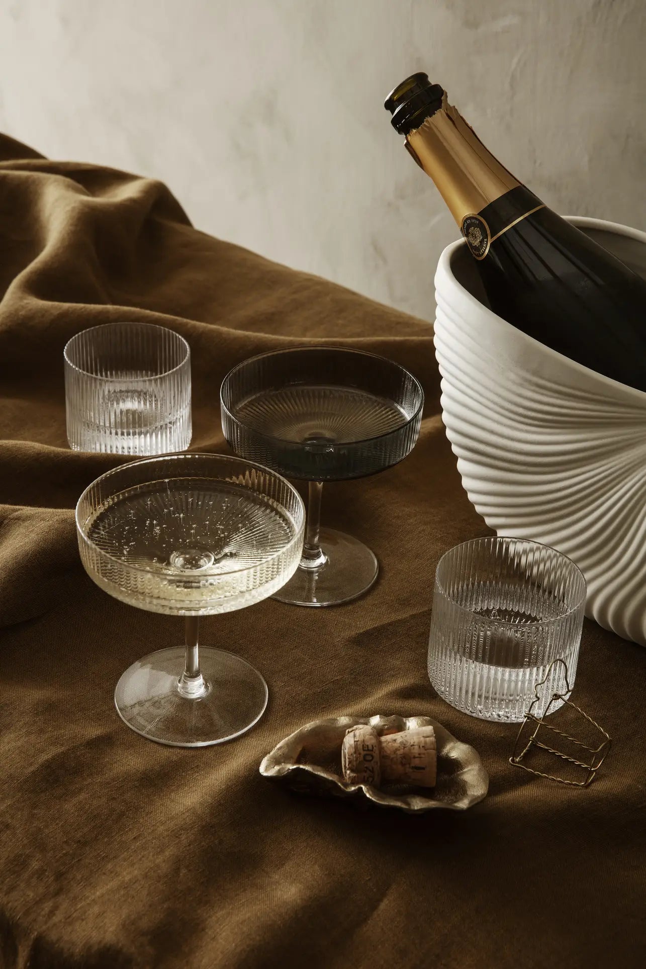 Three elegant coupe glasses and two ribbed tumblers sit beside a white ice bucket with champagne, while the ferm LIVING Shell Pot by ferm LIVING, styled with a cork and wire cage, adds modern design flair to the scene.