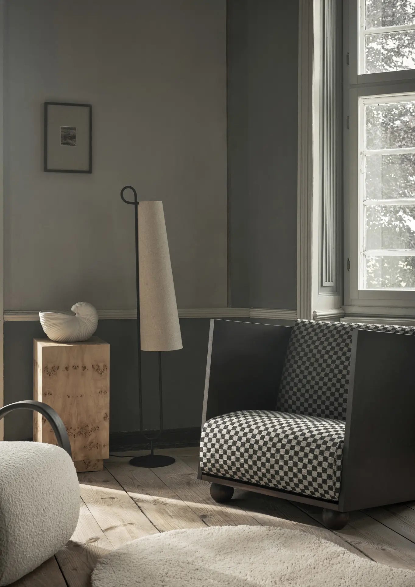 A modern, minimalistic room featuring a checkered armchair, beige textured rug, the ferm LIVING Shell Pot by ferm LIVING with its nautilus shell shape, a slender floor lamp, and a large window flooding the space with natural light.