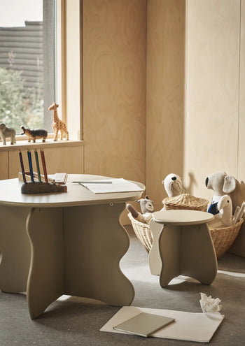 A cozy children’s playroom with a wavy-edged table and a ferm LIVING Slope Stool, both made from FSC-certified MDF, colorful pencils in a holder, open sketchbooks, plush toys in baskets, a toy giraffe on the windowsill, and plenty of natural light.