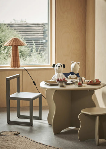A cozy playroom features a round tea table, two stuffed animals, a modern chair, the ferm LIVING Slope Stool, a window with treetop views, and a rattan lamp on the windowsill for organic charm.
