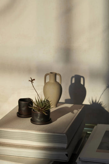 A set of two ferm LIVING Uneru Mini Pots, a dark ceramic cup, and a beige vase rest on a closed book, their soft shadows falling on a light wall bathed in warm sunlight—an inviting plant gift scene.