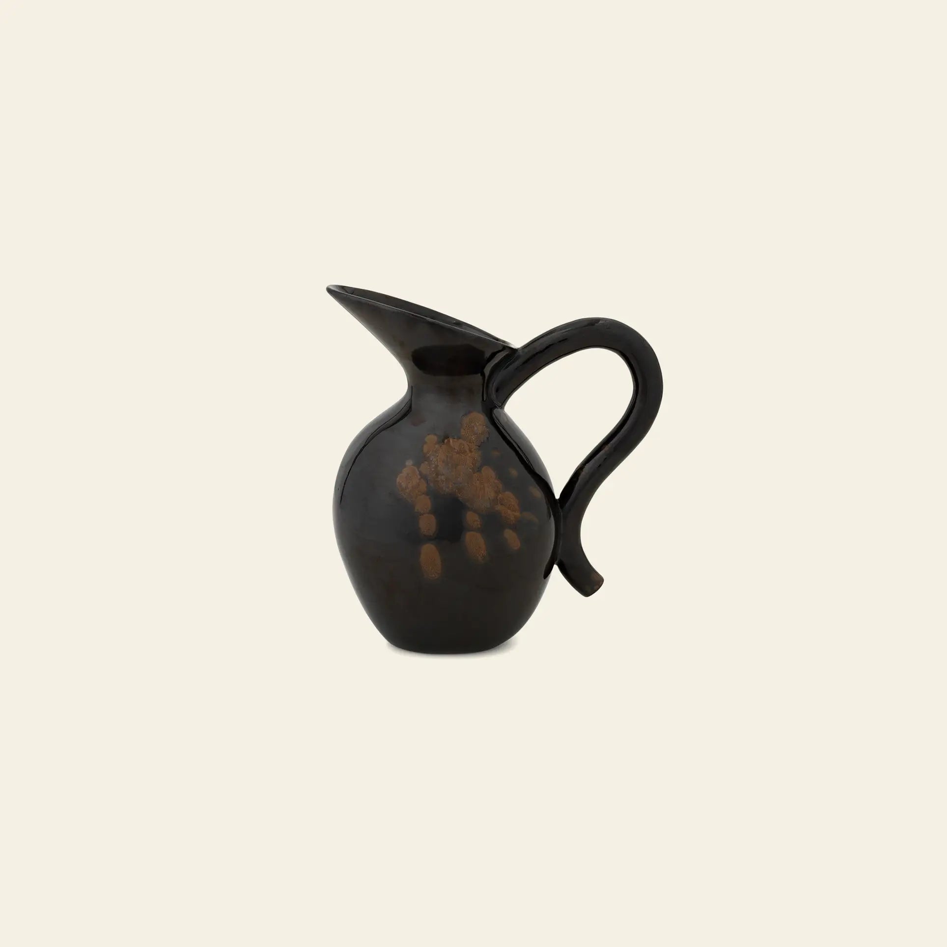The ferm LIVING Verso Jug is a glossy black stoneware piece with a rounded body, wide spout, and curved handle, shown against a plain beige background.