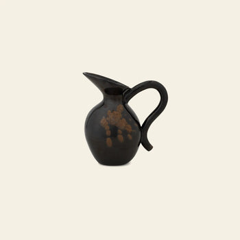 The ferm LIVING Verso Jug is a glossy black stoneware piece with a rounded body, wide spout, and curved handle, shown against a plain beige background.