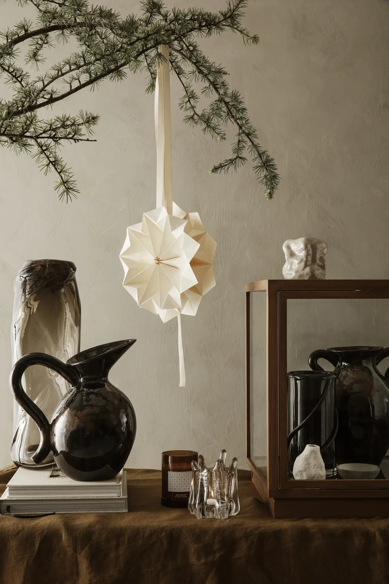 A minimalist still life features the ferm LIVING Verso Jug in dark stoneware, glass objects, and a small sculpture on a brown table. A paper star ornament hangs from a pine branch above, set against neutral-toned walls.
