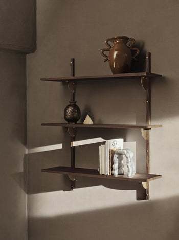 Three dark wooden shelves on a beige wall display a brown vase, small round pottery, the ferm LIVING Verso Table Vase with its organic silhouette, several standing books, and a geometric object. Warm light gives the scene a contemporary touch.