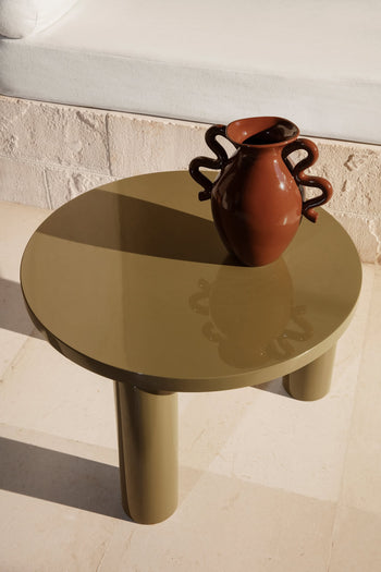 A glossy, round olive-green table with cylindrical legs and a modern edge displays the ferm LIVING Verso Table Vase, featuring sculptural wavy handles. The table is on a light stone floor beside a white cushioned seat.