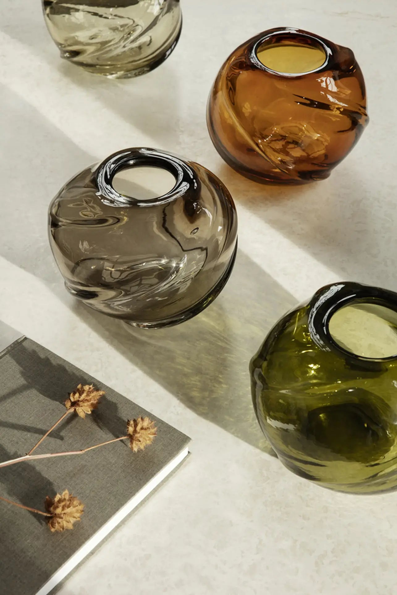 Four round, mouth-blown ferm LIVING Water Swirl Vases in brown, amber, olive green, and smoky gray sit on a light surface by a closed book with dried flowers on its cover, casting soft shadows—each a handcrafted piece.