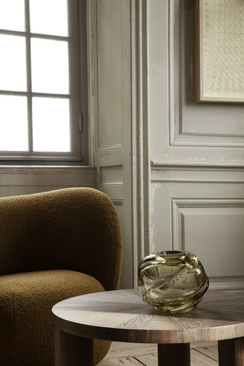 A modern, textured brown armchair sits next to a round wooden table topped with the ferm LIVING Water Swirl Vase, Round, by ferm LIVING, set against ornate gray-paneled walls and a tall window.