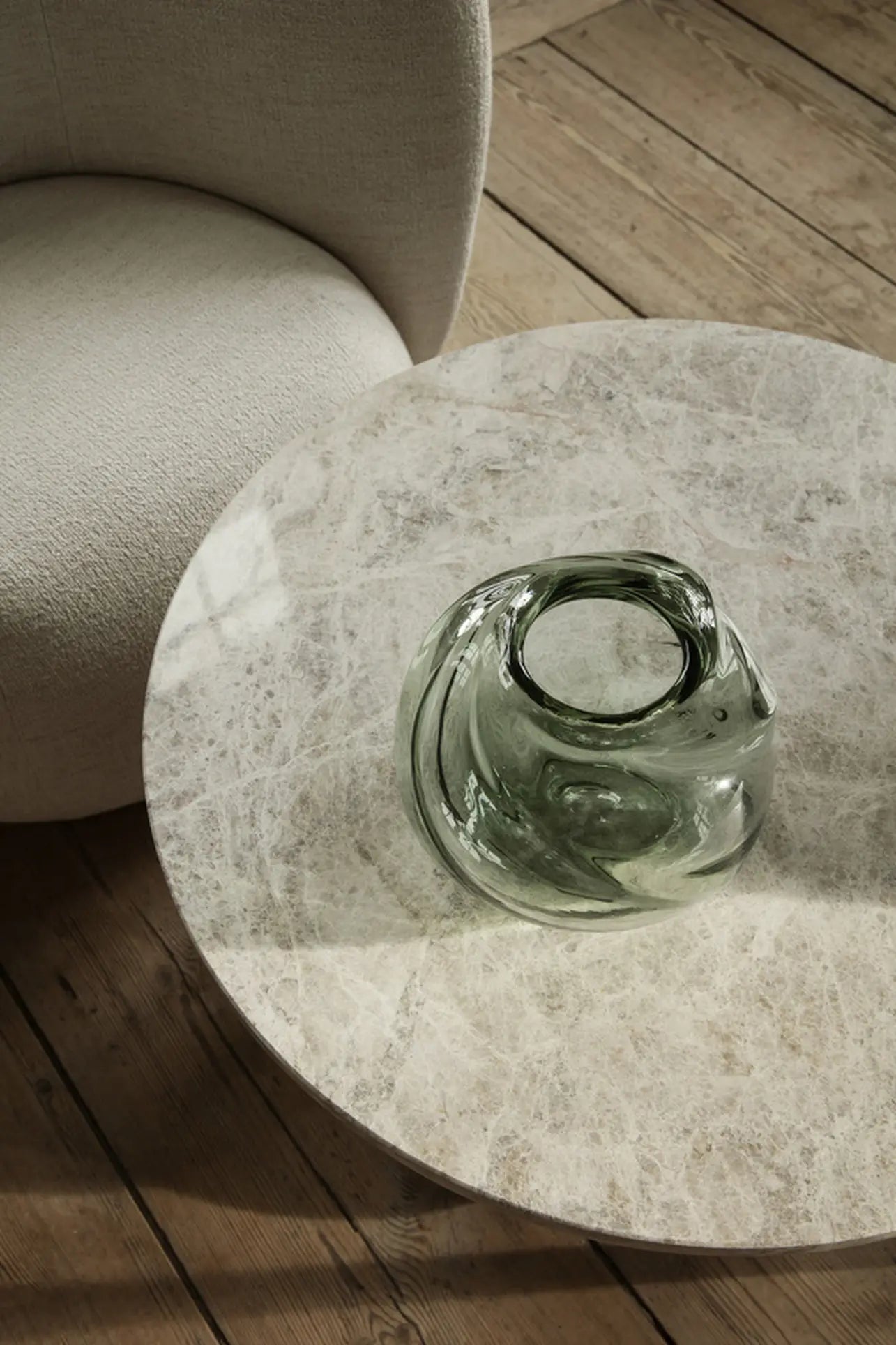 A round marble table with the sculptural ferm LIVING Water Swirl Vase, Round sits beside a light beige upholstered chair on a wooden floor, where natural light casts soft shadows over the handcrafted scene.