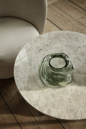 A round marble table with the sculptural ferm LIVING Water Swirl Vase, Round sits beside a light beige upholstered chair on a wooden floor, where natural light casts soft shadows over the handcrafted scene.