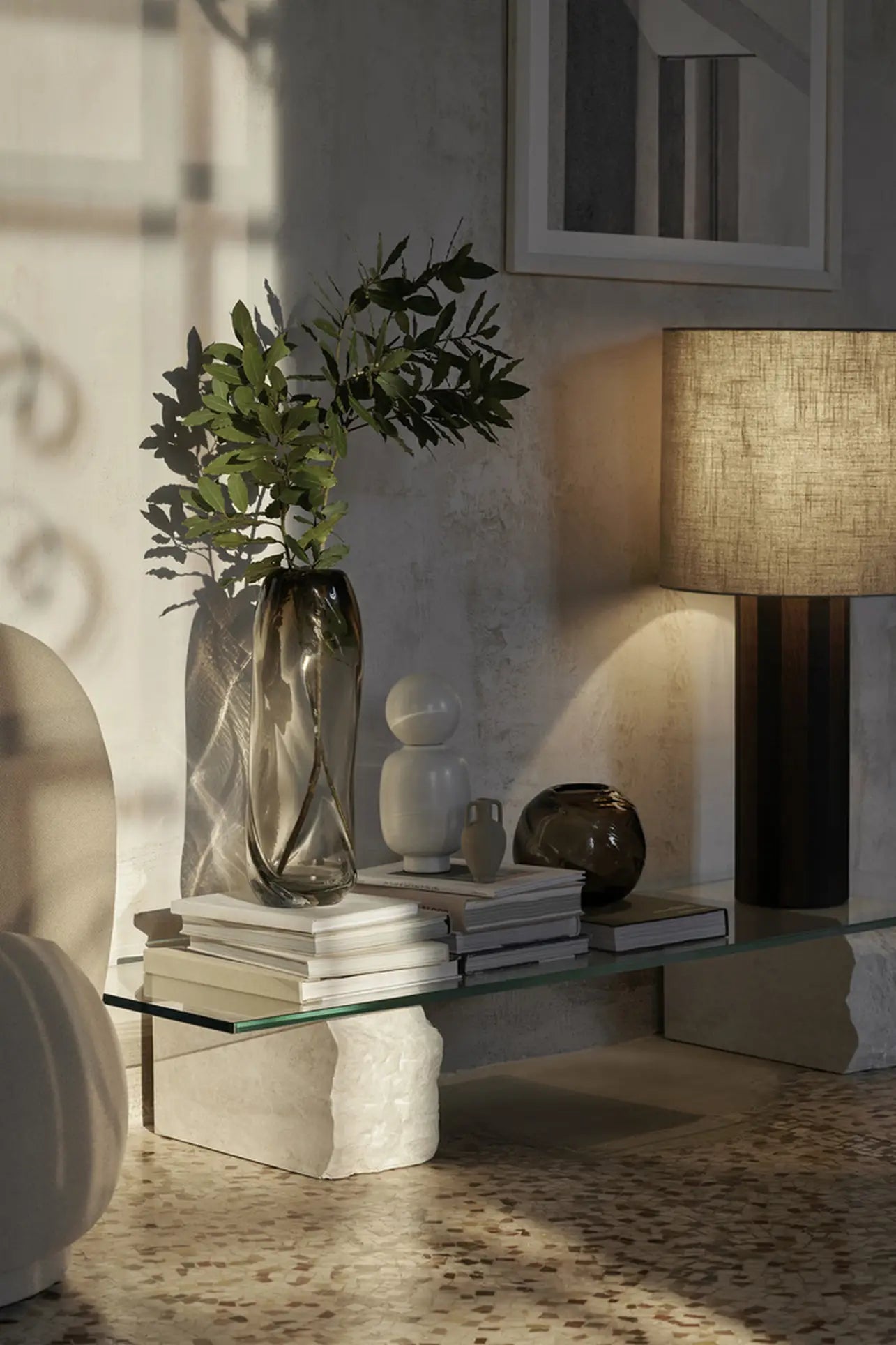 A modern living room corner with a glass coffee table, stacked books, the ferm LIVING Water Swirl Vase, Tall, decorative vases with leafy branches, a sculptural lamp, abstract wall art, and terrazzo flooring—all in soft natural light.
