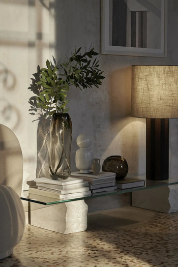 A modern living room corner with a glass coffee table, stacked books, the ferm LIVING Water Swirl Vase, Tall, decorative vases with leafy branches, a sculptural lamp, abstract wall art, and terrazzo flooring—all in soft natural light.