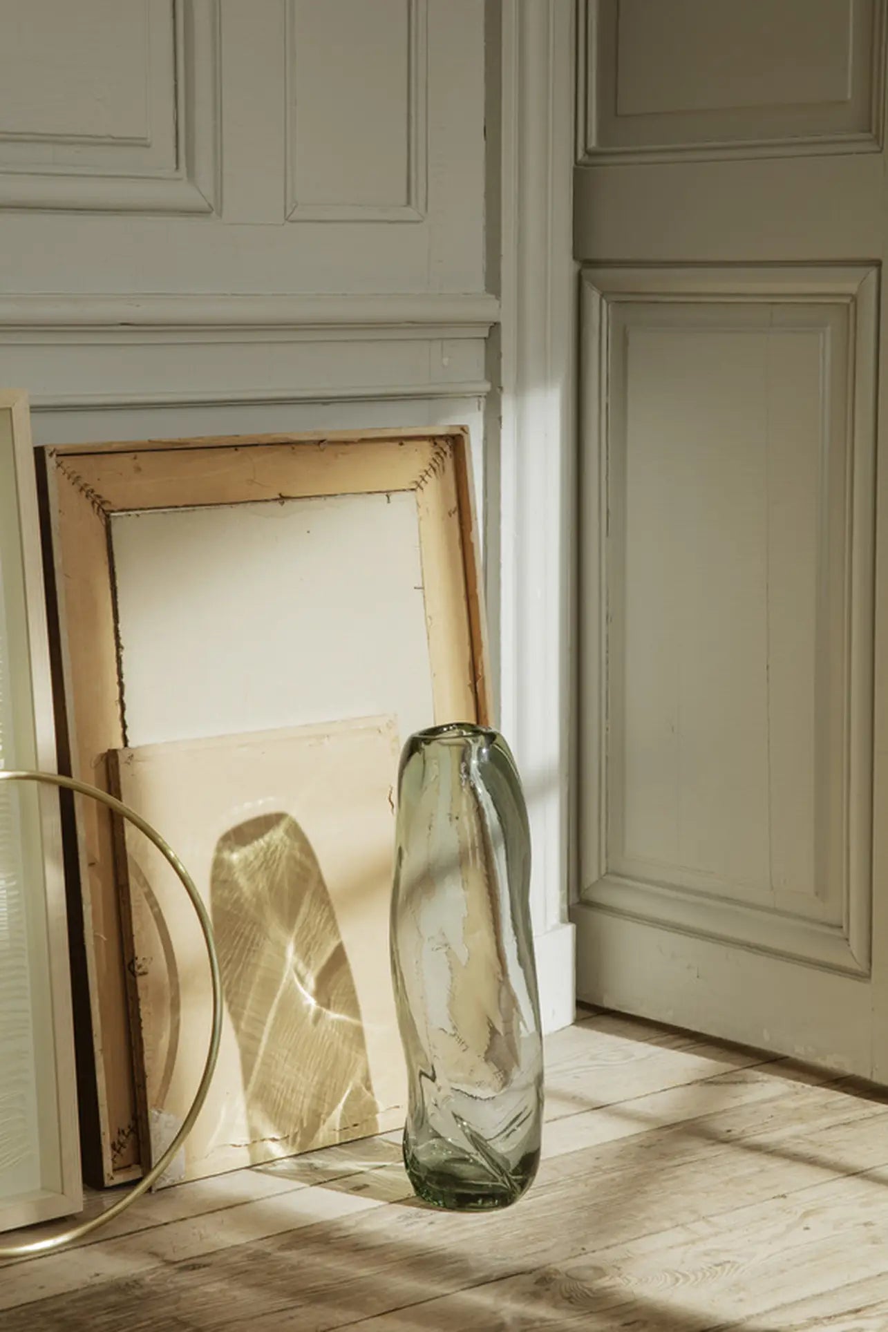 The ferm LIVING Water Swirl Vase, Tall stands on a wooden floor beside two empty stacked frames in a sunlit corner near a white paneled wall and door, with soft shadows evoking a calm, minimalist atmosphere.