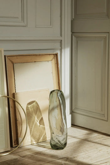 The ferm LIVING Water Swirl Vase, Tall stands on a wooden floor beside two empty stacked frames in a sunlit corner near a white paneled wall and door, with soft shadows evoking a calm, minimalist atmosphere.