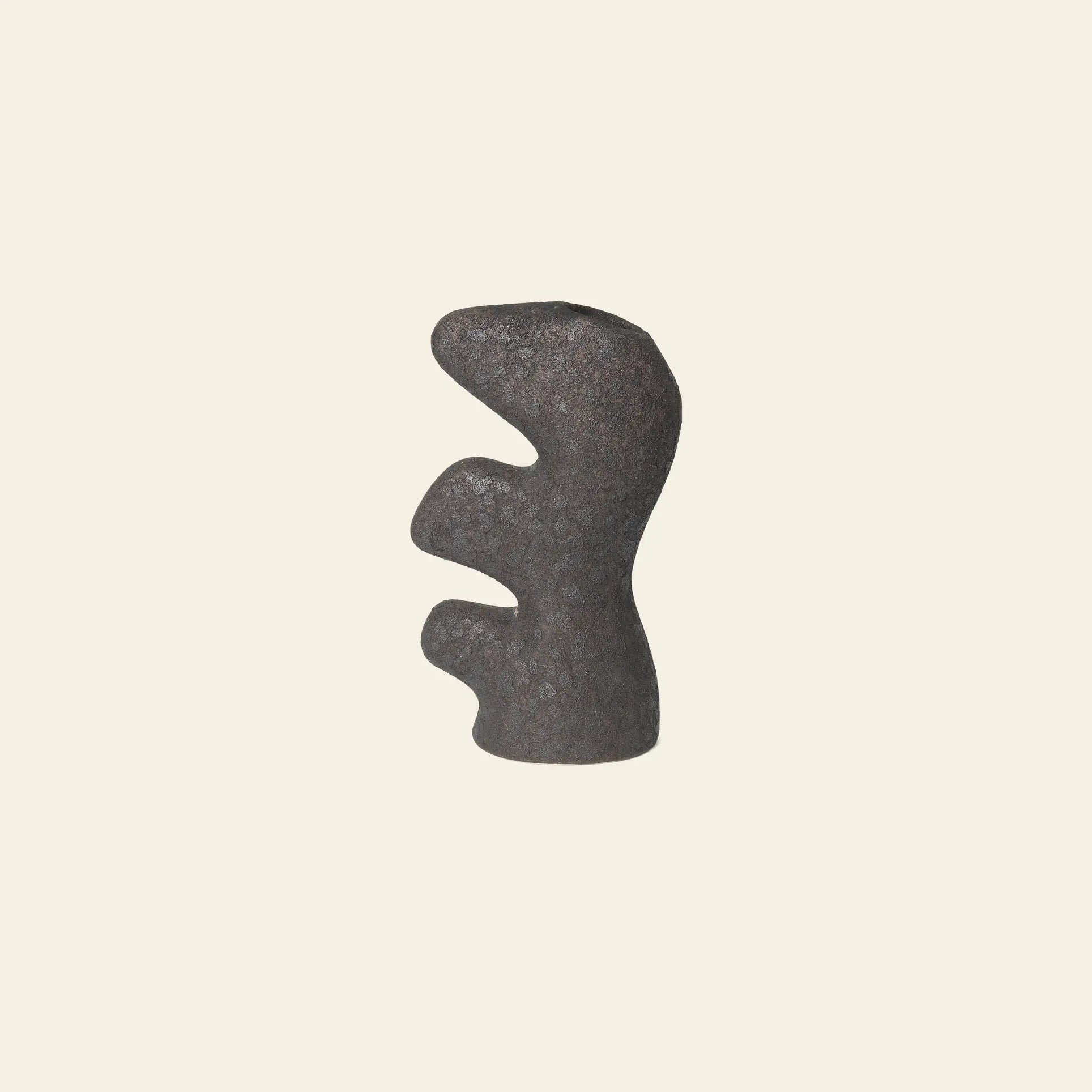 The ferm LIVING Yara Vase, Small by ferm LIVING features a dark, abstract stone-like form with smooth curves and cutouts, inspired by reactive glaze ceramics, shown against a plain beige background.