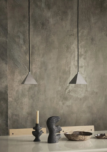 A minimalist dining area features two geometric pendant lights, the ferm LIVING Yara Vase, Small with reactive glaze, a single candle, and decorative bowls on a light table set against a textured gray wall.