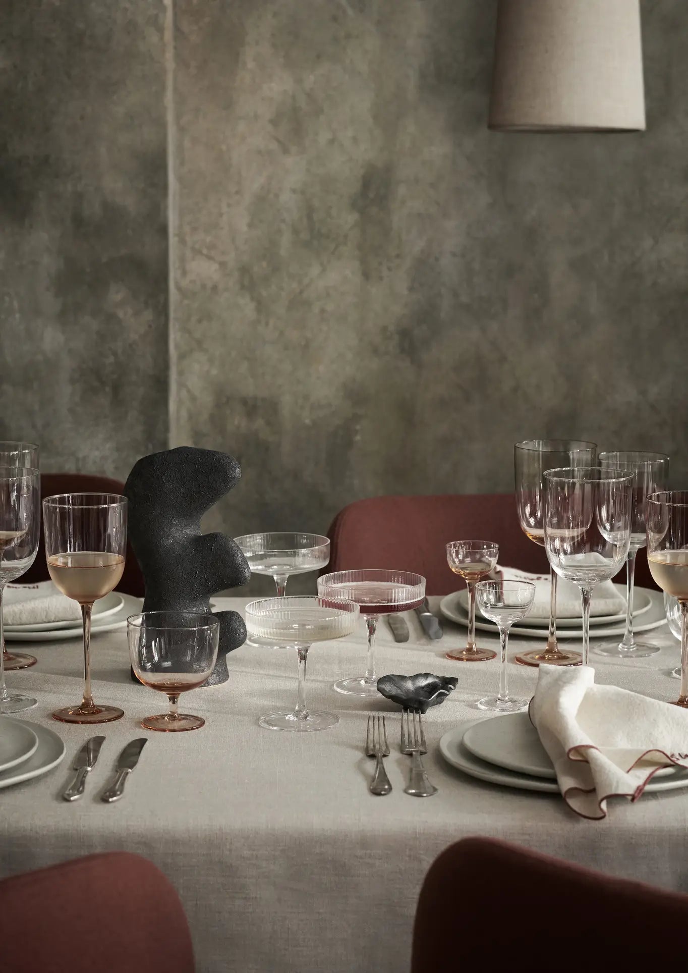 A minimalist dining table is styled with glassware, plates, cutlery, white napkins, a black abstract sculpture centerpiece, and the ferm LIVING Yara Vase, Small by ferm LIVING for a subtle artistic touch against red chairs and a textured gray wall.