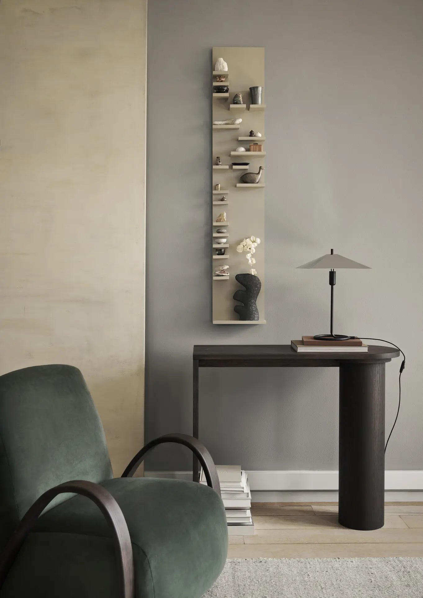 A modern, minimalist room featuring a dark green armchair, a narrow dark table with a lamp and the ferm LIVING Yara Vase, Small by ferm LIVING, small decorative wall shelves, and stacked books on the floor against a gray wall.