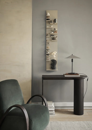 A modern, minimalist room featuring a dark green armchair, a narrow dark table with a lamp and the ferm LIVING Yara Vase, Small by ferm LIVING, small decorative wall shelves, and stacked books on the floor against a gray wall.