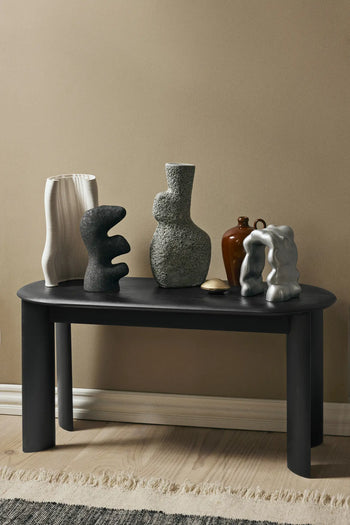 A black table displays five ferm LIVING Yara Vases and sculptures, featuring small ceramic vases with reactive glaze in white, gray, black, and brown, set against a beige wall on a light wood floor near a fringed rug.