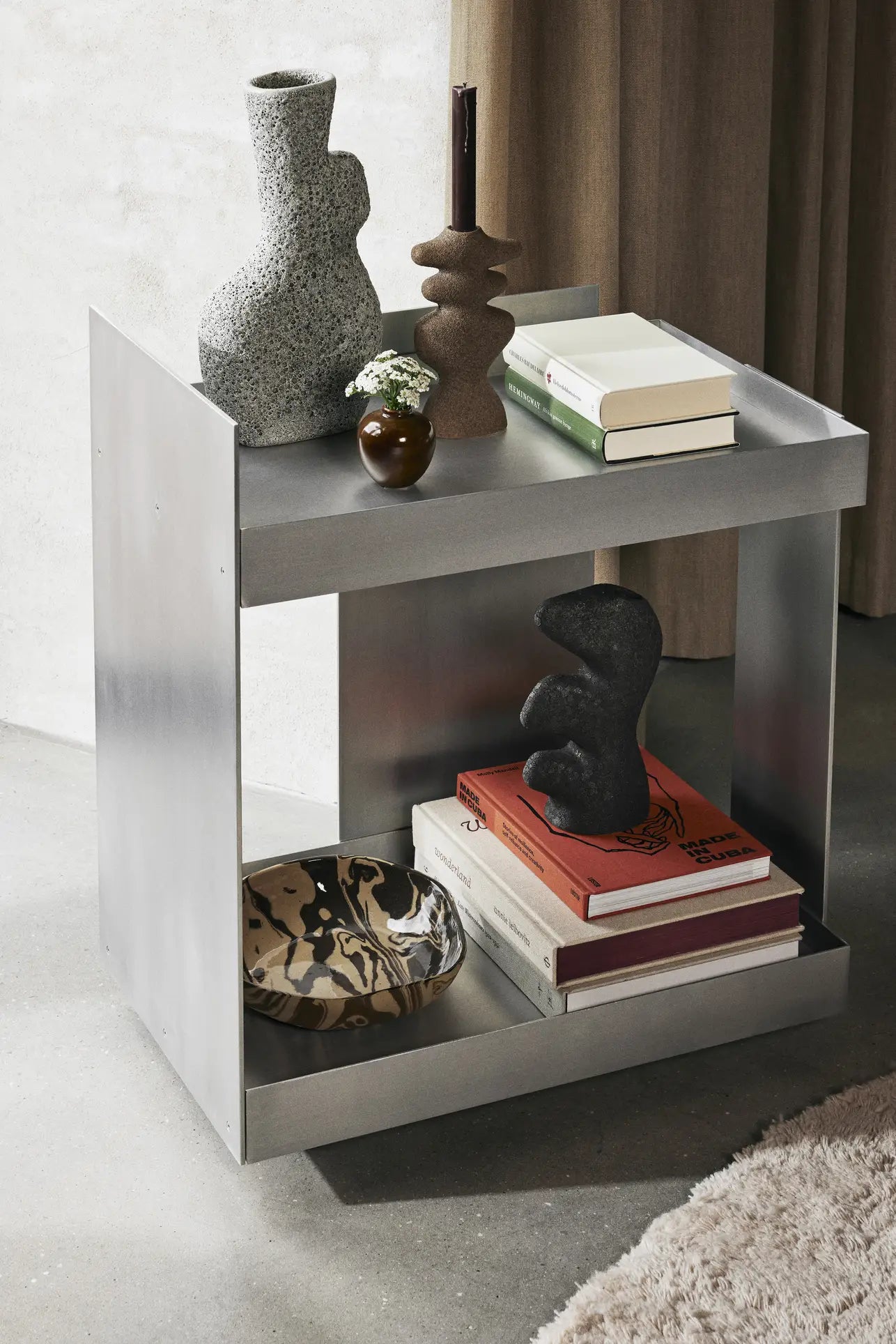 A modern metallic side table displays the ferm LIVING Yara Vase, Small, with reactive glaze, stacked books, a floral arrangement, and a patterned bowl. The scene highlights minimalist decor in neutral tones with artistic accents.