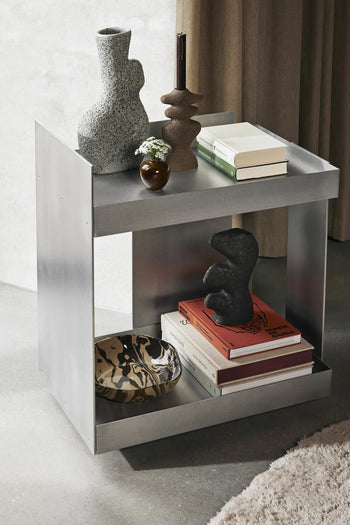 A modern metallic side table displays the ferm LIVING Yara Vase, Small, with reactive glaze, stacked books, a floral arrangement, and a patterned bowl. The scene highlights minimalist decor in neutral tones with artistic accents.