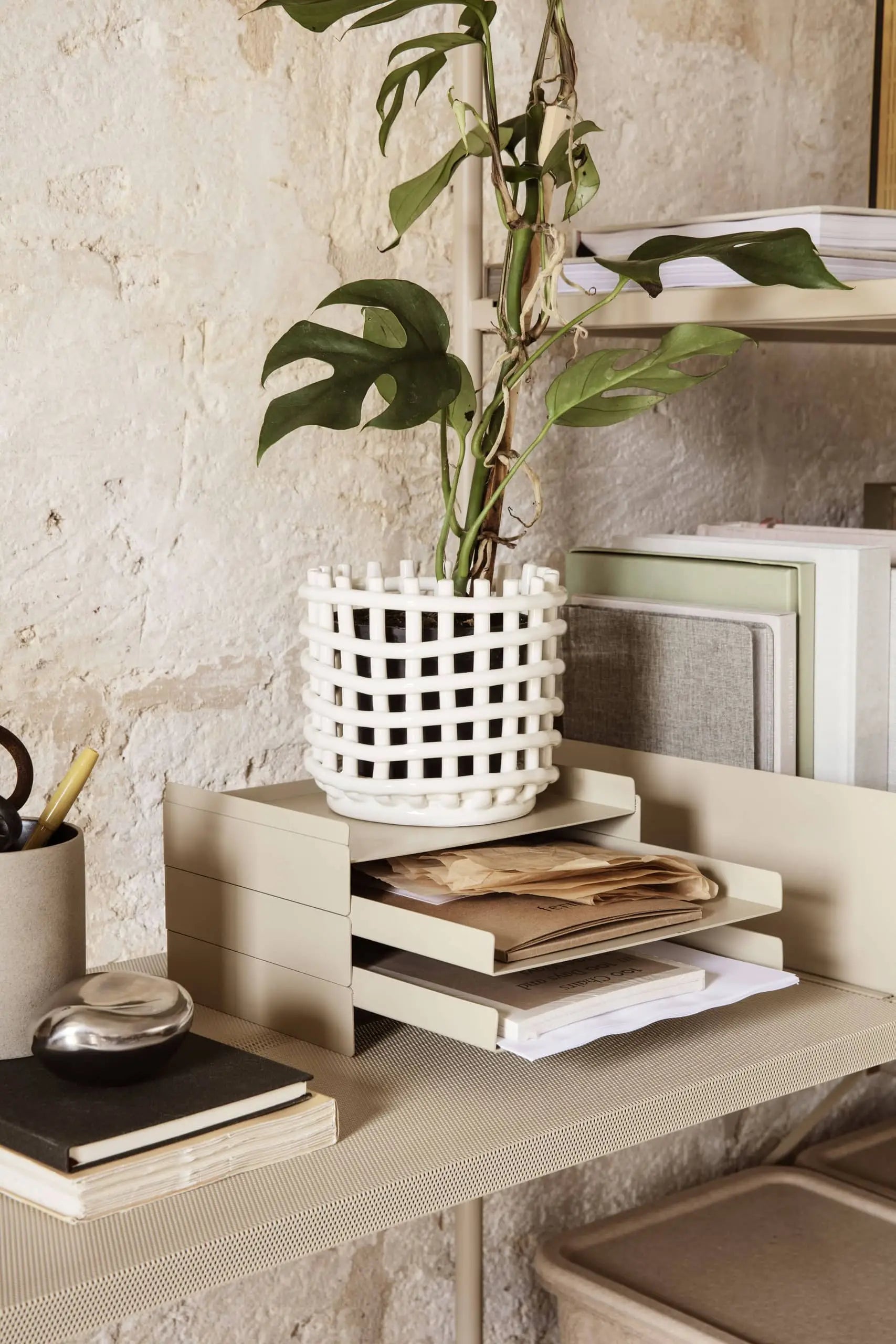 The ferm LIVING 2x2 Organizer by ferm LIVING, paired with a notebook, pen, and a white lattice plant pot, keeps documents tidy on a modern desk set against a textured stone wall.