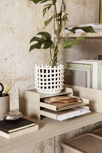 The ferm LIVING 2x2 Organizer by ferm LIVING, paired with a notebook, pen, and a white lattice plant pot, keeps documents tidy on a modern desk set against a textured stone wall.