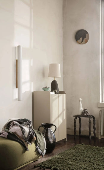 A cozy, modern room with neutral walls, a cabinet topped with a lamp and decor, a small side table, round wall art, a green sofa with blankets, a black vase, and the ferm LIVING Amass Long Pile Mat from ferm LIVING. Sunlight streams in.