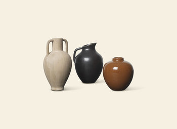 Three ceramic vases sit side by side on a beige background: a tall beige vase with two handles, a black jug with handle and spout, and the ferm LIVING Ary Mini Vase L in round brown textured glazed stoneware.