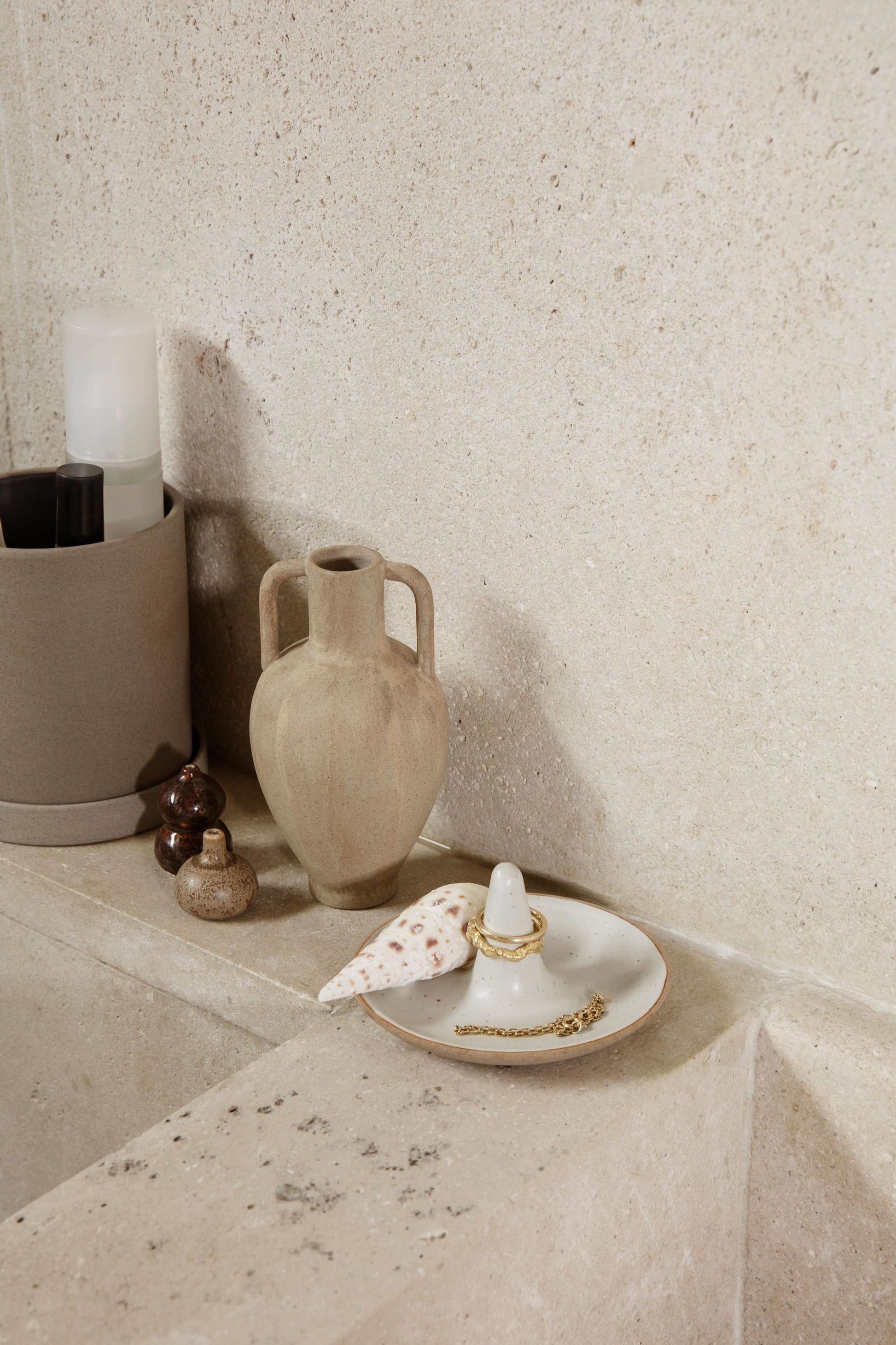 A beige stone bathroom counter features the ferm LIVING Ary Mini Vase L in textured glazed stoneware, a round container with bottles, a plate of gold jewelry, a ring holder, a seashell, and two small brown Greek-inspired ornaments.