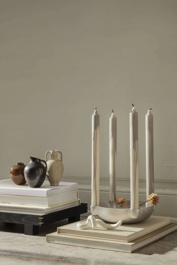 A minimalist display with four beige marbled candles in a silver holder, flanked by the ferm LIVING Ary Mini Vase M and small brown ceramics on stacked books, all set against a neutral beige wall.