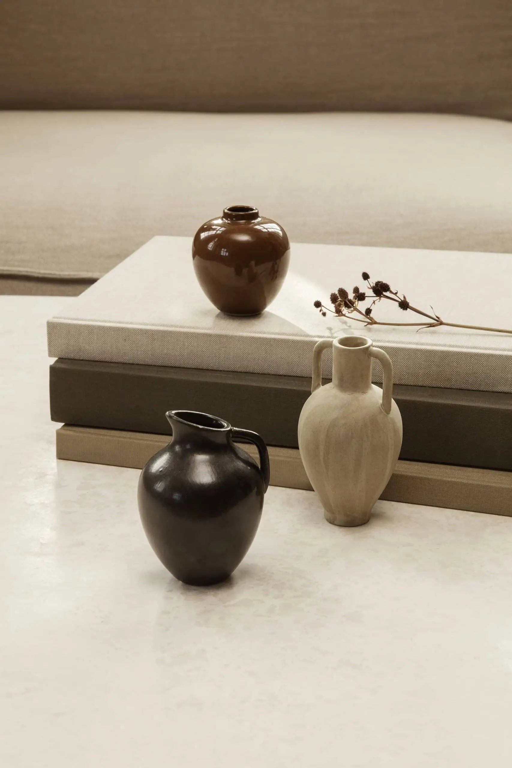 The ferm LIVING Ary Mini Vase S by ferm LIVING sits with dried flowers on and beside stacked neutral books, adding stylish decor to a light textured surface in front of a beige sofa. Its brown, black, and beige hues recall classic Greek vases.
