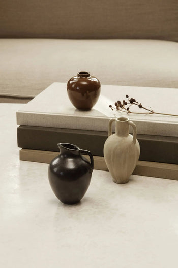 The ferm LIVING Ary Mini Vase S by ferm LIVING sits with dried flowers on and beside stacked neutral books, adding stylish decor to a light textured surface in front of a beige sofa. Its brown, black, and beige hues recall classic Greek vases.