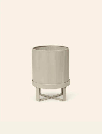 A beige, cylindrical ferm LIVING Bau Pot, Small with vertical grooves rests on a matching X-shaped stand against an off-white background, echoing the clean lines of Bauhaus architecture.