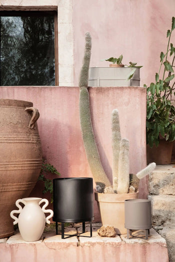 A tall cactus in the ferm LIVING Bau Pot, Small is displayed on stone steps among decorative planters, vases, and a large clay jar, set against a pink wall with leafy plants in the background.