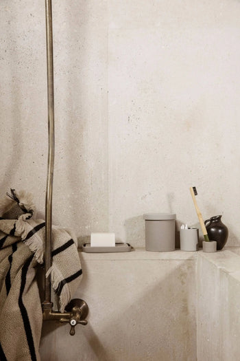 A minimalist bathroom scene with neutral walls, a striped towel, soap dish, lidded container, and a bamboo toothbrush in the ferm LIVING Bon Accessories Toothbrush Holder beside a small black vase.