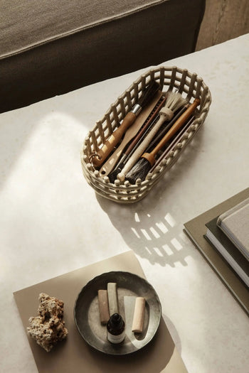 The ferm LIVING Ceramic Basket Oval displays brushes and wooden tools on a white tabletop. Surrounding it are books, a gray ceramic plate with chalk, a bottle, and sponges—all by ferm LIVING, in soft neutral colors with contemporary style.