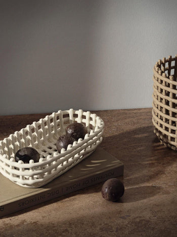 The ferm LIVING Ceramic Basket Oval by ferm LIVING, filled with three avocados, rests on a closed book. Nearby, another avocado and a larger basket add modern style and practical storage in natural light against a plain wall.