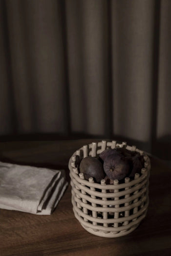 The ferm LIVING Ceramic Basket, Small, by ferm LIVING, filled with dark figs sits on a wooden surface beside a folded beige cloth and soft brown curtains—a stylish and elegant table storage solution.