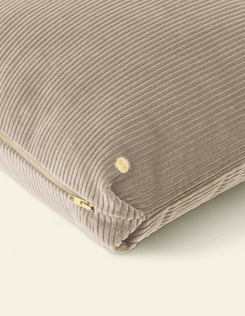 A close-up of the ferm LIVING Corduroy Cushion in beige with a corduroy texture, gold zipper, and a small round gold tag, set against a light background.