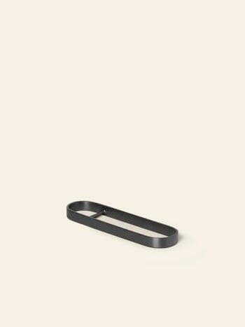 The ferm LIVING Fein Bottle Opener by ferm LIVING, featuring a minimalist black metal design with an oval shape and low edge, is displayed on a plain light beige background—perfect for a contemporary kitchen.
