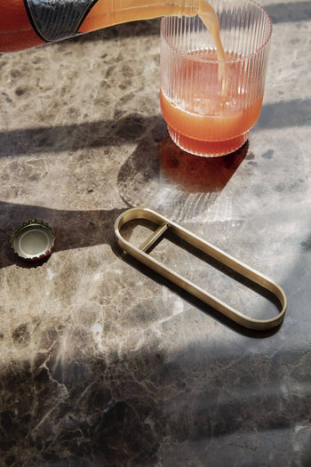 A glass of pinkish-orange drink sits on a marble table in a modern kitchen, next to the brass ferm LIVING Fein Bottle Opener and a green bottle cap. Sunlight casts soft shadows, emphasizing the minimalist style.